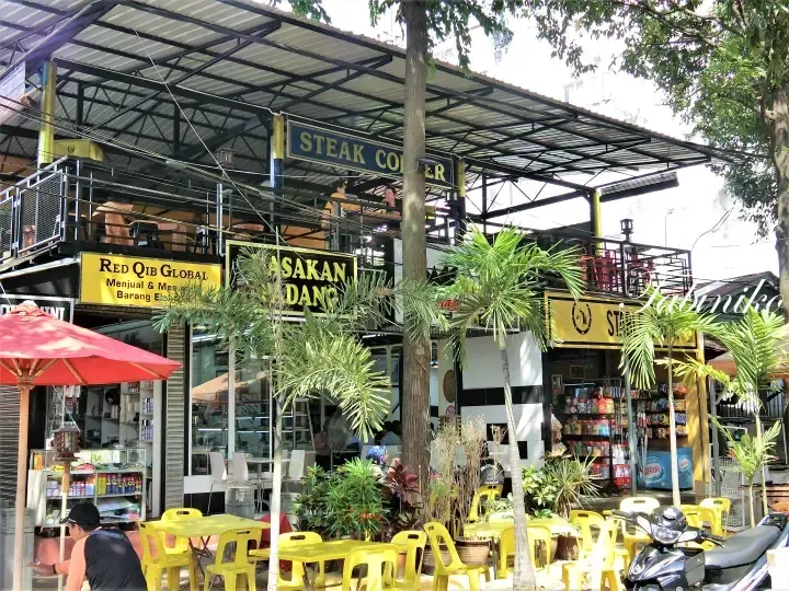 Outdoor dining area with yellow tables and chairs in front of a building featuring a Steak Corner sign and a Red Qib Global shop.