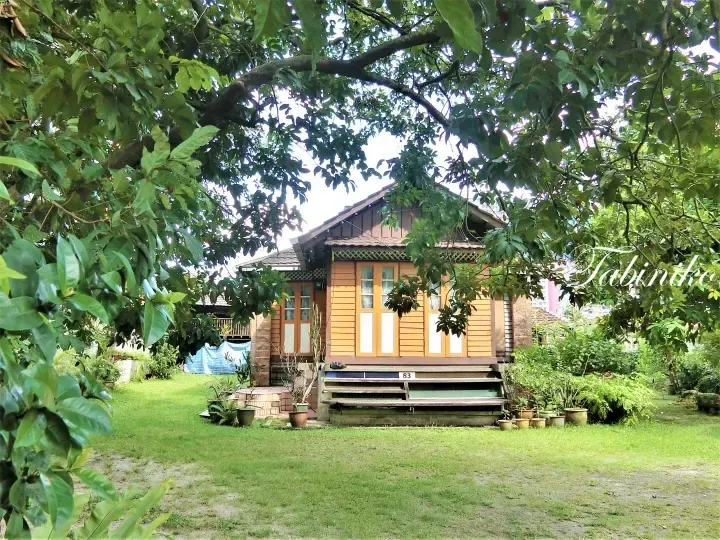 Traditional wooden house with yellow siding and number 83, surrounded by lush greenery and trees in a garden.