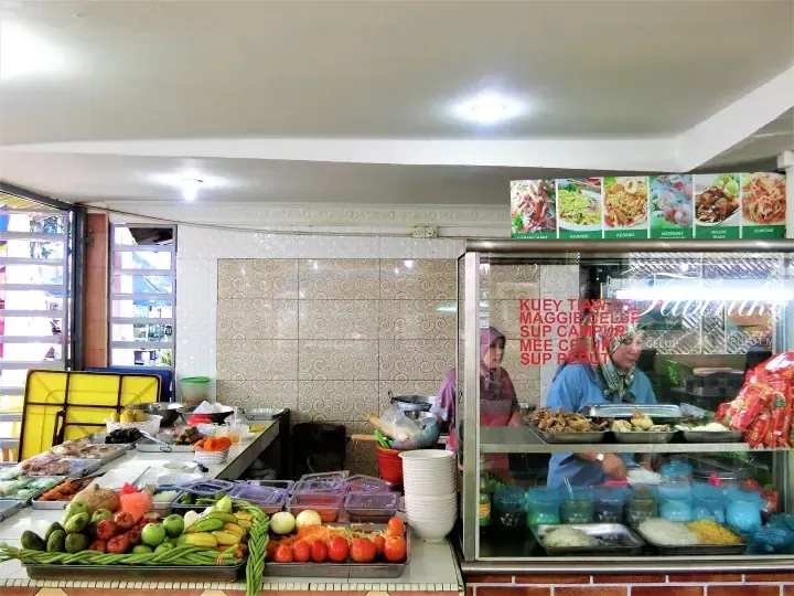 Food stall counter displaying fresh vegetables and menu items like Kuey Tiaw Celup and Maggie Celup in a Malaysian eatery.