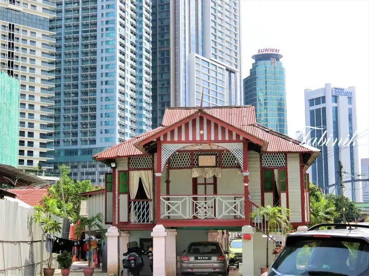 Traditional Malay wooden house with red roof in front of modern skyscrapers, including Sunway and Menara Safuan towers.