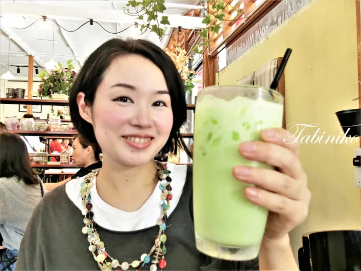 カフェで緑色のアイスドリンクを手に笑顔を見せる女性。背景に植物と棚があり、右下に「Tabiniko」のロゴが確認できる。