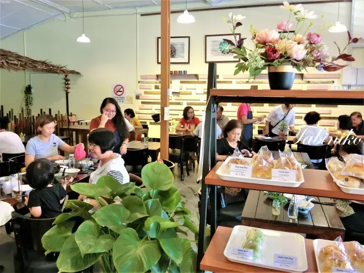 Customers dining inside a cozy cafe with a display shelf featuring packaged buns like Hog Dog Bun and Tuna Bun.