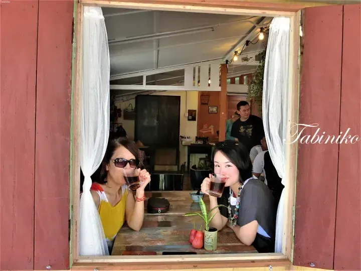 Two women enjoy drinks through a rustic wooden window at a cafe in Malaysia.
