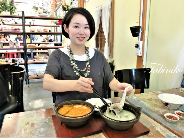 レストランのテーブルで、2つのスープのボウルに向かって箸を持った女性が笑顔で写っています。