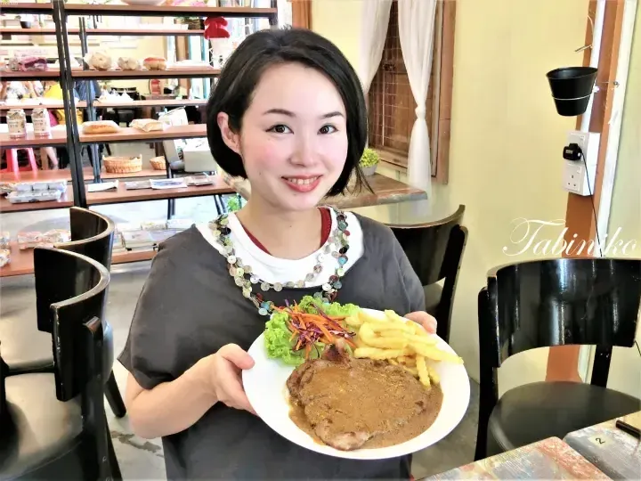 Smiling woman holding a plate of steak with gravy, fries, and salad in a cafe setting.