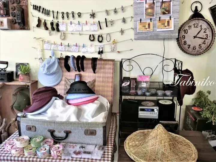 Vintage shop interior displaying hats, jewelry, and accessories on a suitcase and wall, with a large clock and &lsquo;Tabiniko&rsquo; watermark.