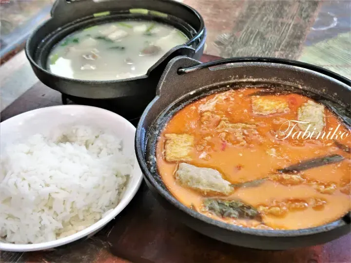 Two cast iron pots with white and red soups served alongside a bowl of white rice on a wooden table.