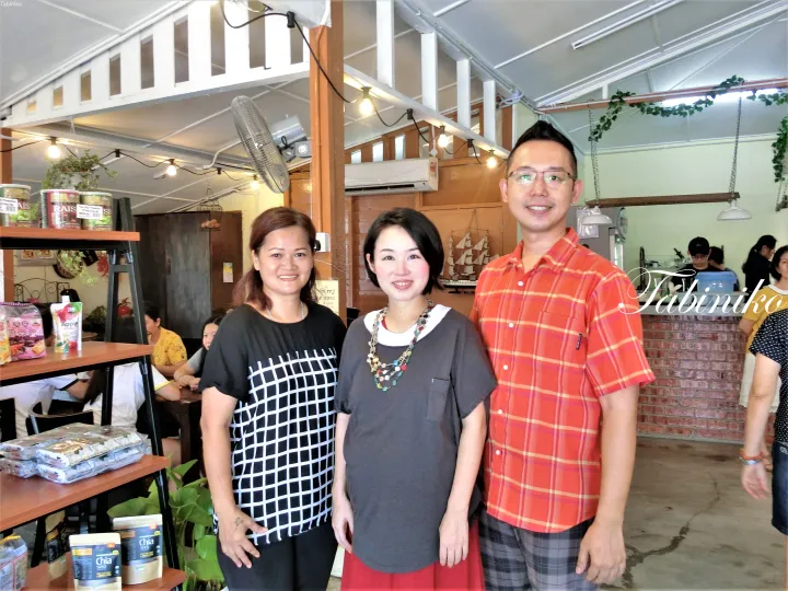 Chia Seedなどの商品が並ぶ木製棚のある、居心地の良いカフェでポーズをとる3人。