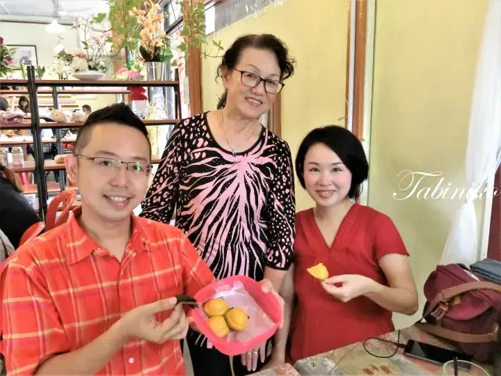 Three people smiling in a cafe, holding a pink bowl of yellow kuih and a piece of food.