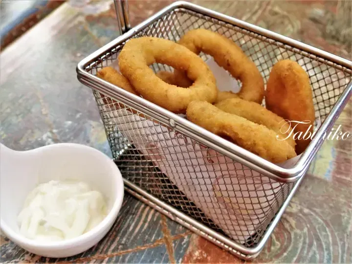 Golden fried onion rings served in a wire basket with a side of creamy white dipping sauce.