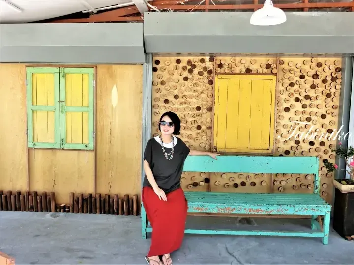 Woman in sunglasses and red skirt posing on a turquoise bench against a rustic wooden wall with colorful shutters.