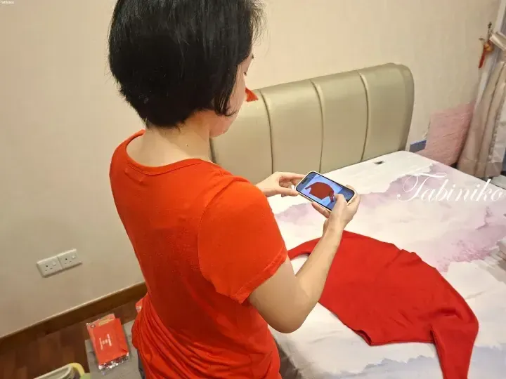 Woman in red shirt taking a photo of a red garment on a bed with a smartphone.