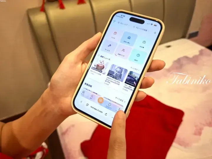 A hand holding a smartphone displaying the tsunagu.it.com app interface with Japanese text and news headlines for Malaysia.
