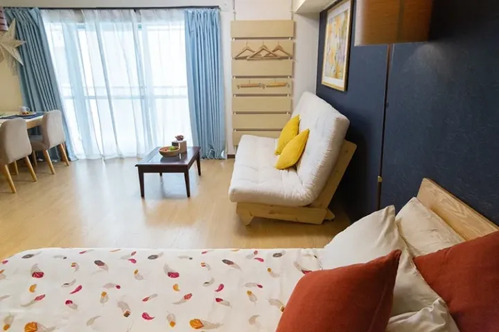 Bright studio apartment with a feather-patterned bed, yellow cushion sofa, and large window with blue curtains.