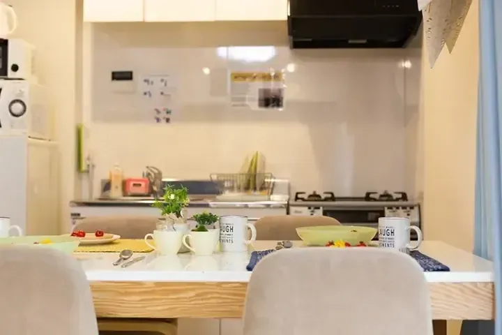調理器具や食器が並ぶ明るいダイニングテーブルと、その奥のモダンなキッチンが映し出された室内風景。