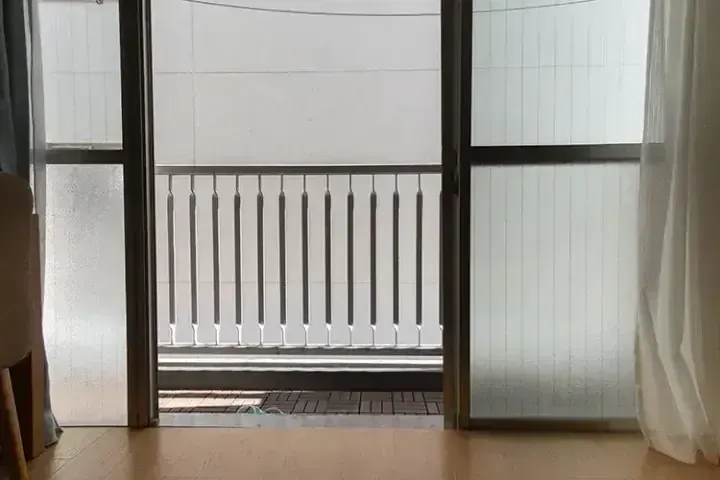 從鋪有木質地板的房間透過敞開的滑門，看到帶有金屬欄杆和磨砂玻璃面板的陽台。