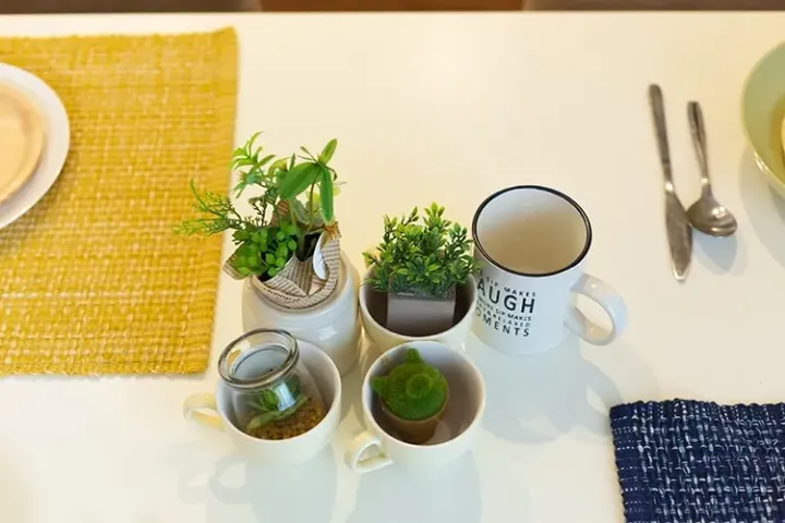 Minimalist table setting featuring small potted plants, a mug with text, and cutlery on a white surface with yellow and blue placemats.