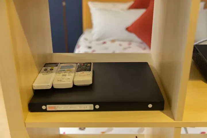 Three remote controls resting on a black binder inside a light wooden shelf unit in a hotel room.