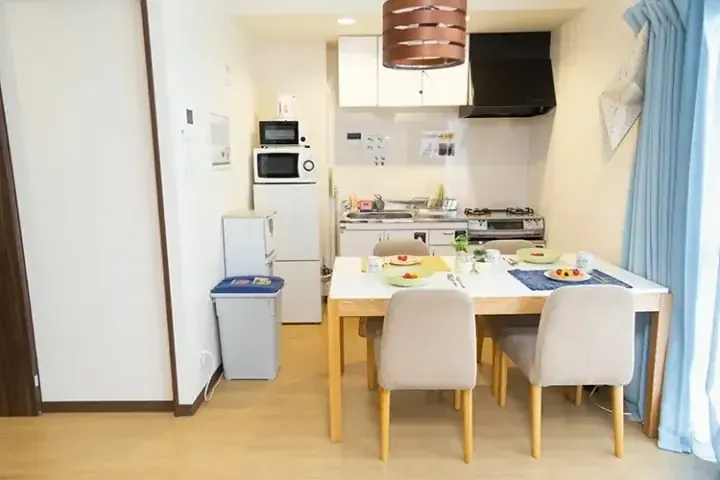 白い家電と銅色のペンダントライトが特徴のコンパクトなキッチンに隣接する、木製テーブルと椅子のある明るいダイニングエリア。