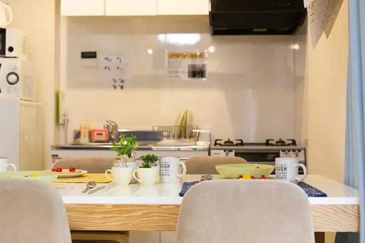 Bright dining table with mugs and plants overlooking a modern kitchen with a stove and microwave in a Malaysian home.