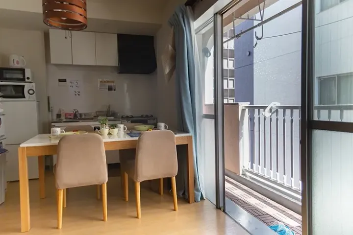 Modern apartment dining area with wooden table, beige chairs, kitchenette, and sliding glass door opening to a balcony.