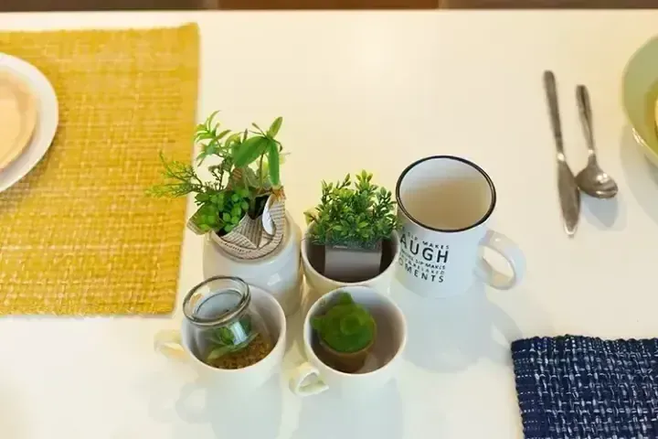 白色桌面上擺放著小型盆栽、印有文字的馬克杯、餐具以及黃藍色餐墊的簡約餐桌佈置。