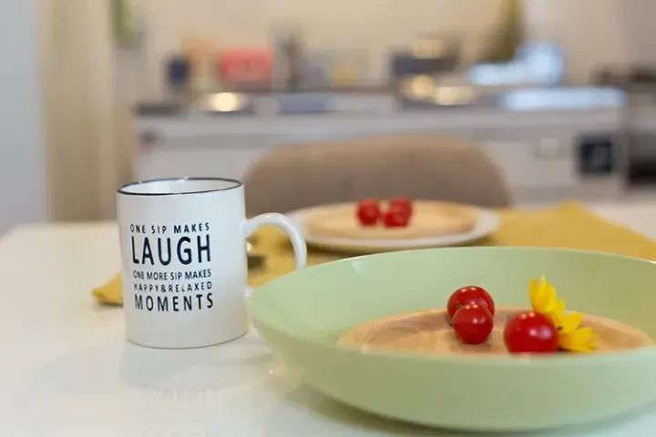 A white mug with inspirational text sits beside a bowl of soup garnished with cherry tomatoes and a yellow flower on a kitchen table.