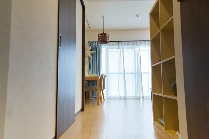Bright hallway with wooden floors leading to a dining area with a table, chairs, and a window with sheer curtains.