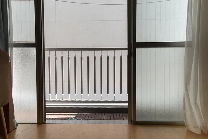 View from a wooden-floored room through open sliding doors onto a balcony with a metal railing and frosted glass panels.