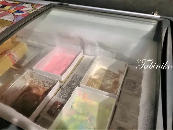 霜が降りたガラス蓋付き冷凍庫に並ぶ、白い容器に入った様々なフレーバーのジェラート。