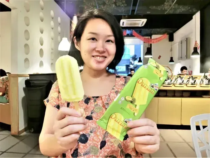 Woman smiling while holding a durian ice cream bar and its green packaging inside a modern cafe.