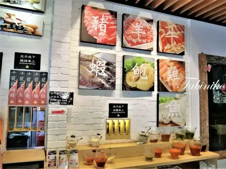 Restaurant wall displaying illuminated panels of meat and seafood options like pork, lamb, beef, prawn, abalone, chicken, and fish above a condiment bar.