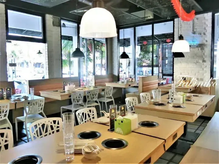 Bright dining area with wooden tables featuring built-in hotpot burners, white chairs, and large windows overlooking the street.