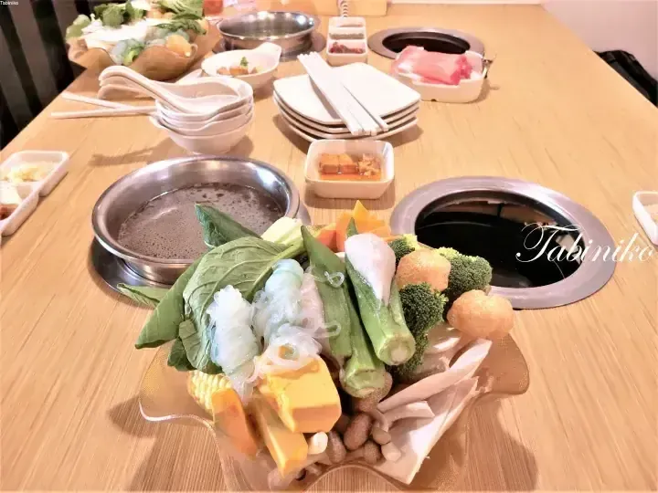 A table set for hot pot with a central grill, surrounded by fresh vegetables, tofu, noodles, and raw meat.