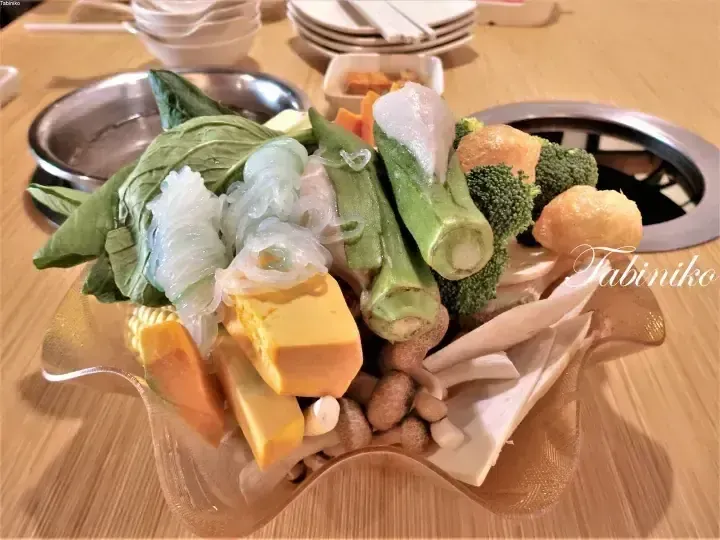 A colorful assortment of fresh vegetables, tofu, and mushrooms arranged in a glass bowl ready for hot pot.