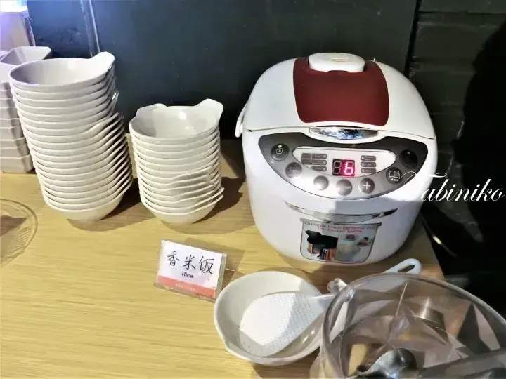 A rice cooker and stacks of white bowls on a wooden counter with a sign reading &ldquo;Rice&rdquo;.