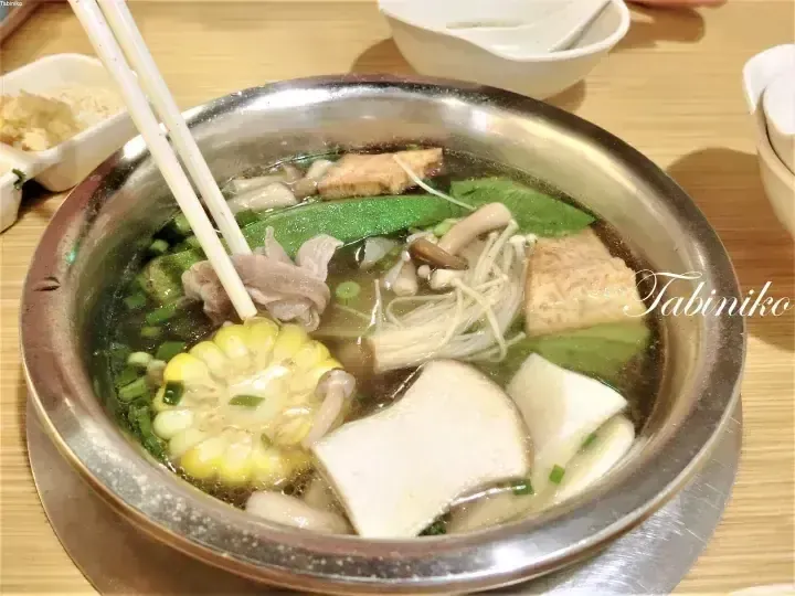 A steaming metal bowl filled with hot pot ingredients including corn, mushrooms, tofu, and meat, with chopsticks resting on the edge.