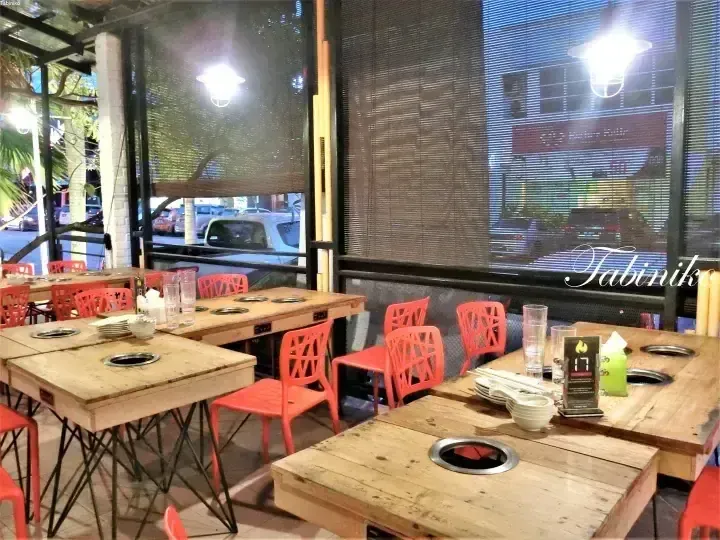 Wooden tables with built-in grills and orange chairs at a restaurant interior with large windows overlooking a street.