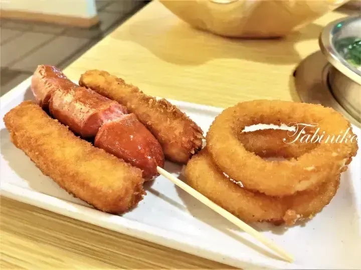 Fried sausages, corn dogs, and onion rings served on a white rectangular plate with a wooden skewer.