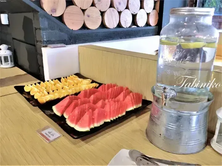 Sliced watermelon and orange arranged on a buffet tray alongside a glass water dispenser at a dining area.