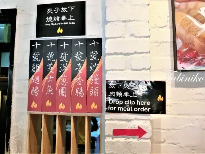 Menu signs at a Malaysian BBQ restaurant showing items like chicken wings and sausages with instructions to drop clips for orders.