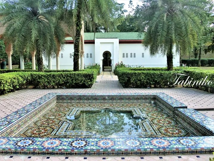 Colorful mosaic fountain in a courtyard with palm trees and a white building featuring arched doorways.