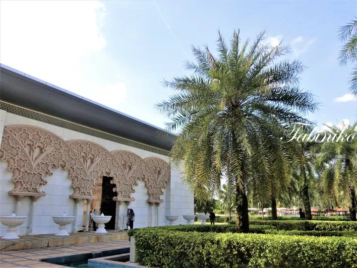 Ornate Moorish arches and white fountains at the entrance of Putrajaya Mosque surrounded by palm trees.