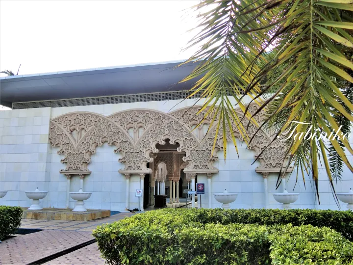 Ornate Moorish arches and white marble fountains at the entrance of a mosque in Malaysia, framed by palm leaves.