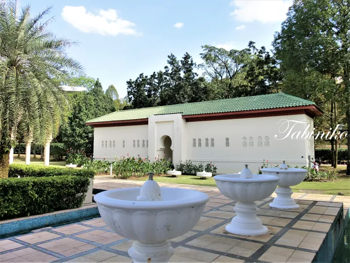 White mosque with green roof and arched entrance surrounded by lush gardens and decorative fountains.