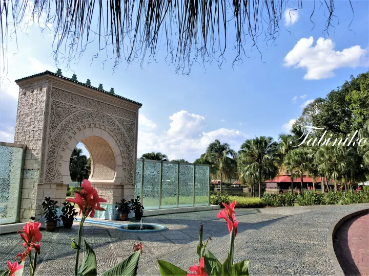Ornate archway at the entrance of a tropical resort with palm trees and red flowers under a blue sky.