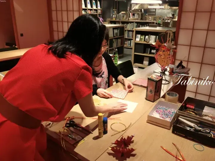 赤いドレスの女性が、本棚のある温かい部屋で、木製のテーブルで折り紙のクラフトをゲストに教える。
