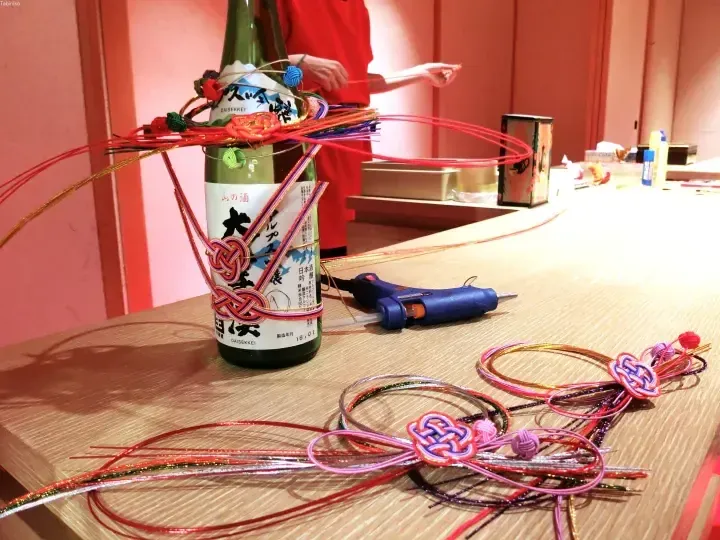 木工テーブルの上で、色とりどりの紐とホットボンドガンで日本酒の瓶を飾り付けしている様子。