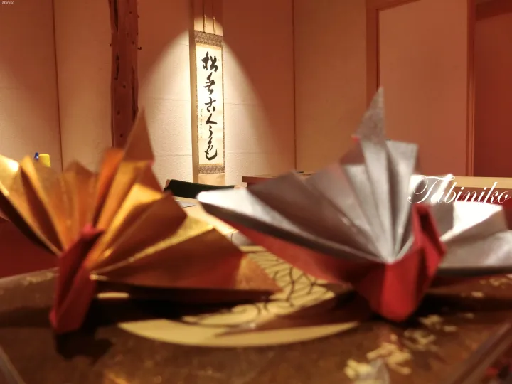 Golden and silver origami cranes on a table with a calligraphy scroll in the background.