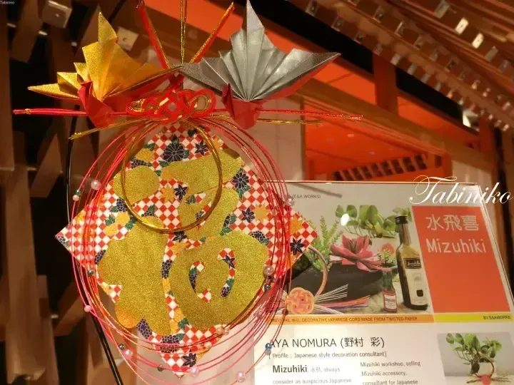 由紅色繩結裝飾的金色和銀色紙花組成的日本傳統水飛喜裝飾品，旁邊放有說明牌。